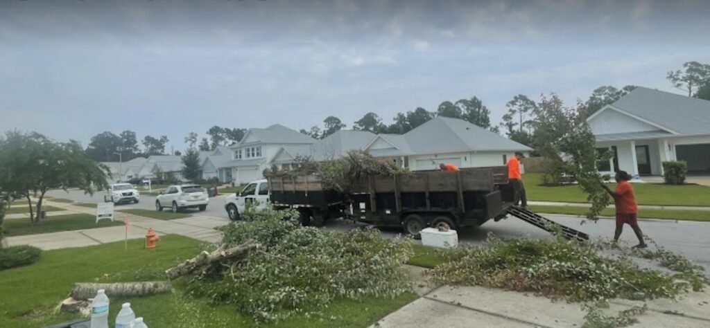 Cornerstone crew in orange hi-vis loading cut tree limbs and brush into two dump trailers on a residential street in Northwest Florida